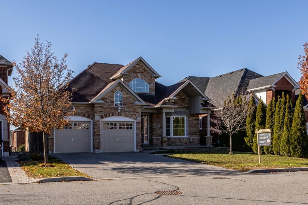 A charming stone-faced house with a landscaped yard, featuring two garages, under a clear blue sky, with a "For Sale" sign.