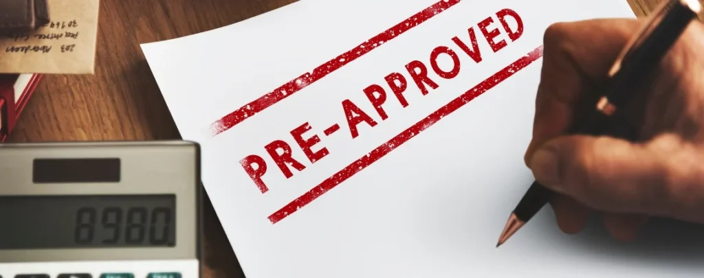 A hand holding a pen is writing on a document marked "PRE-APPROVED," alongside a calculator and books on a desk.