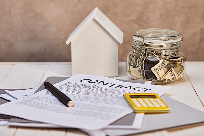A house model beside a contract, a calculator, and a jar filled with cash on a wooden table, symbolizing real estate transactions.