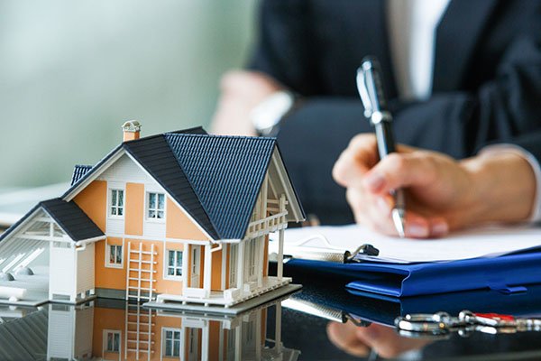 A miniature house model rests on a desk, with a person in a suit signing documents in the background, illustrating real estate transactions.